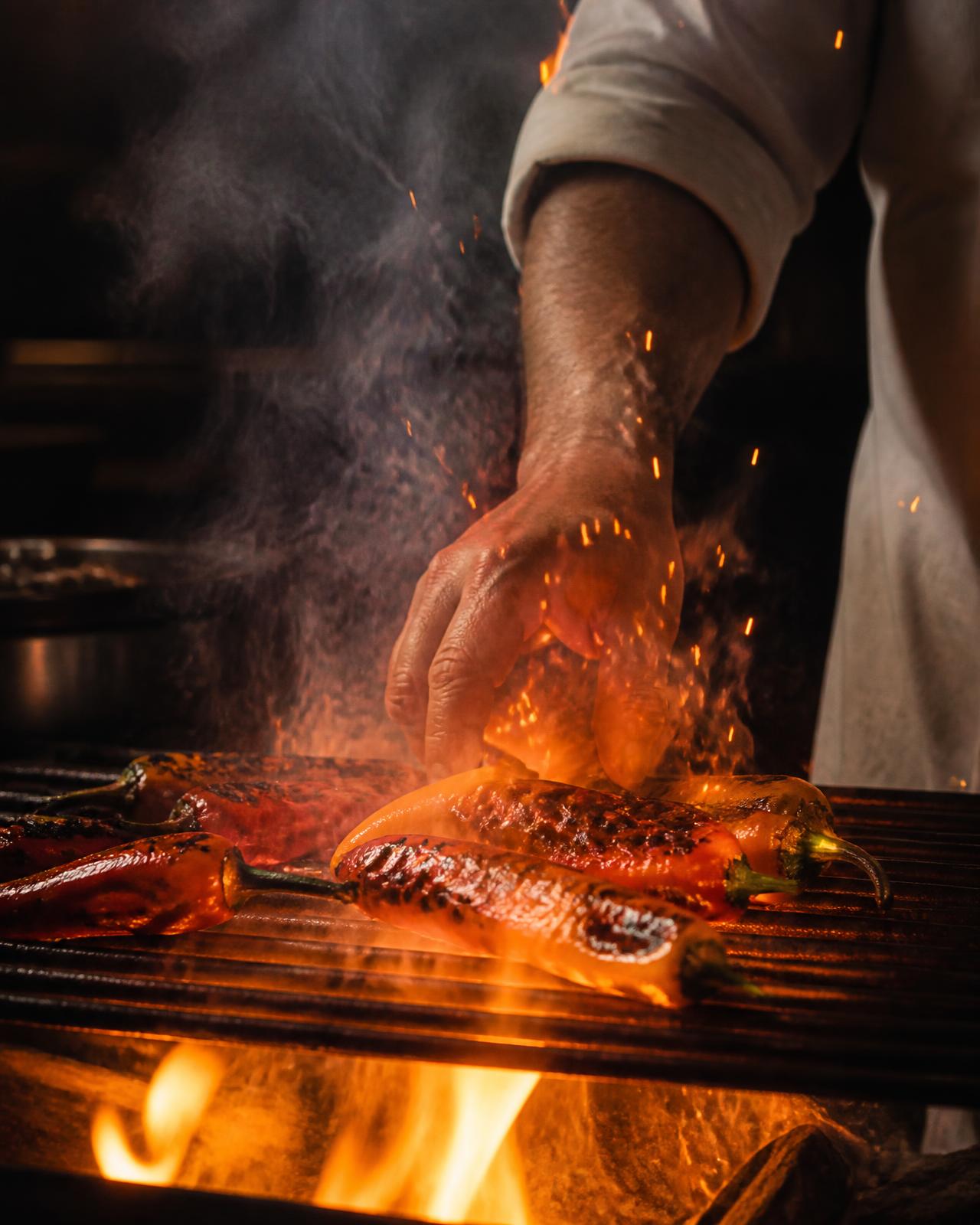 Chef's hand tending peppers over the flames